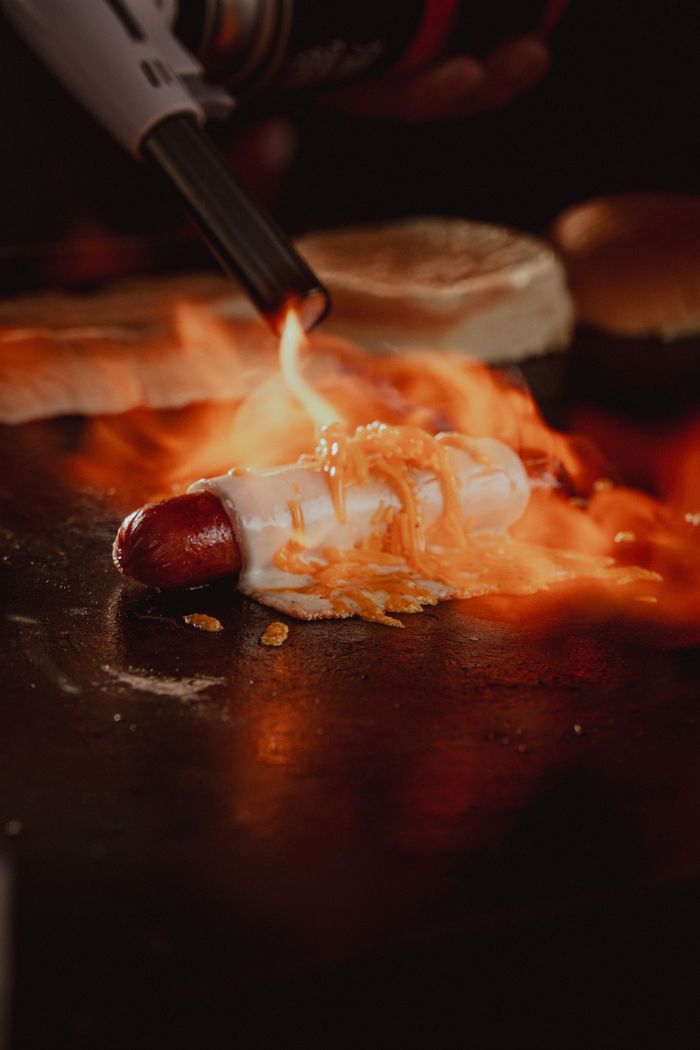 about-03 A delicious gourmet hot dog is being grilled with a torch flame, giving it a perfect crisp finish.