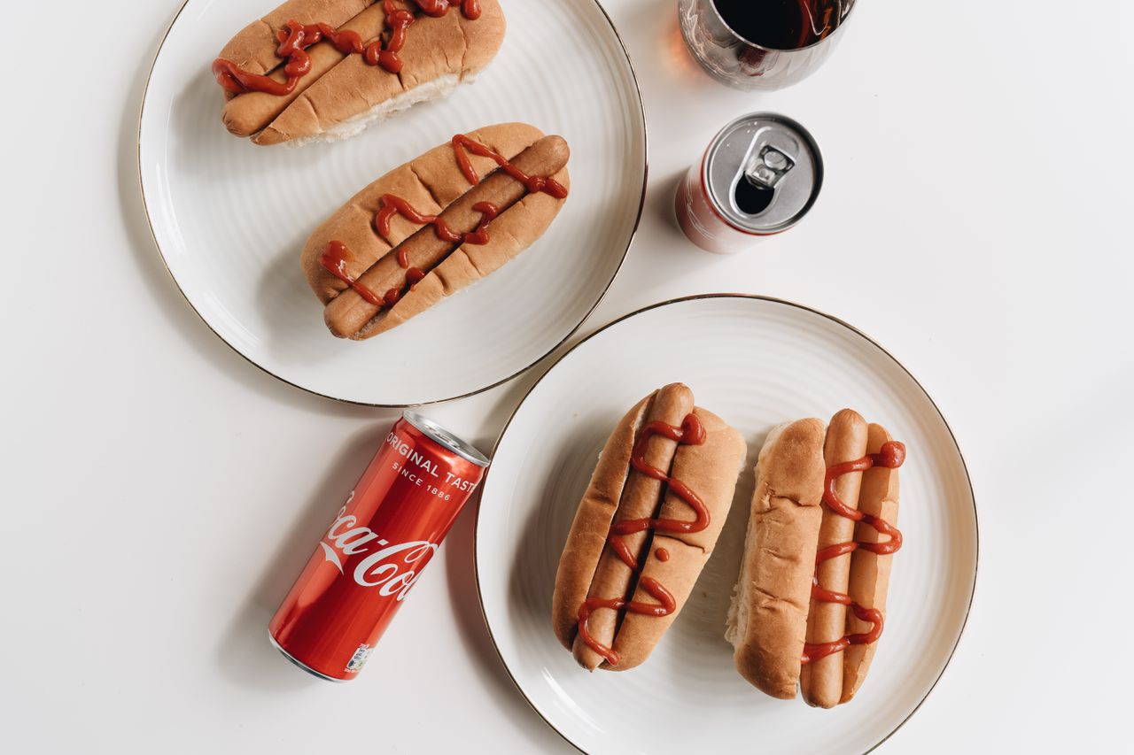 product-04 Delicious hot dogs with ketchup paired with Coca Cola cans on plates in a flat lay setting.