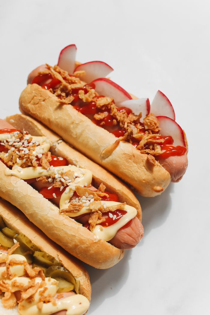 Close-up of three mouth-watering gourmet hotdogs on a white background, garnished with various toppings.