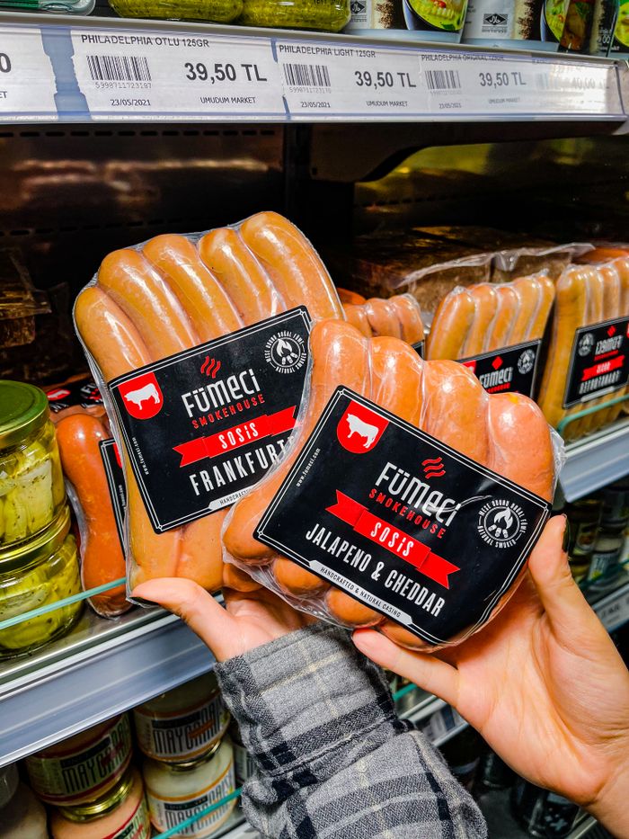 Close-up of hands holding packaged sausages in a grocery store refrigerator section.